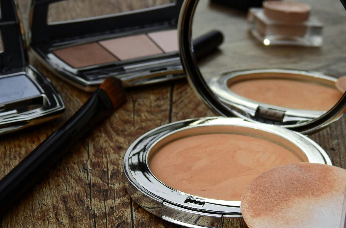 Close-up of makeup tools including compact, brush, and mirror on a rustic wooden surface.